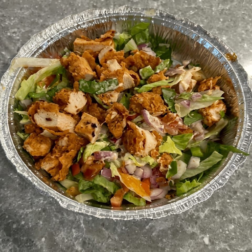 Aluminum bowl filled with green salad, crispy fried chicken pieces, chopped vegetables, and dressing on a gray countertop.
