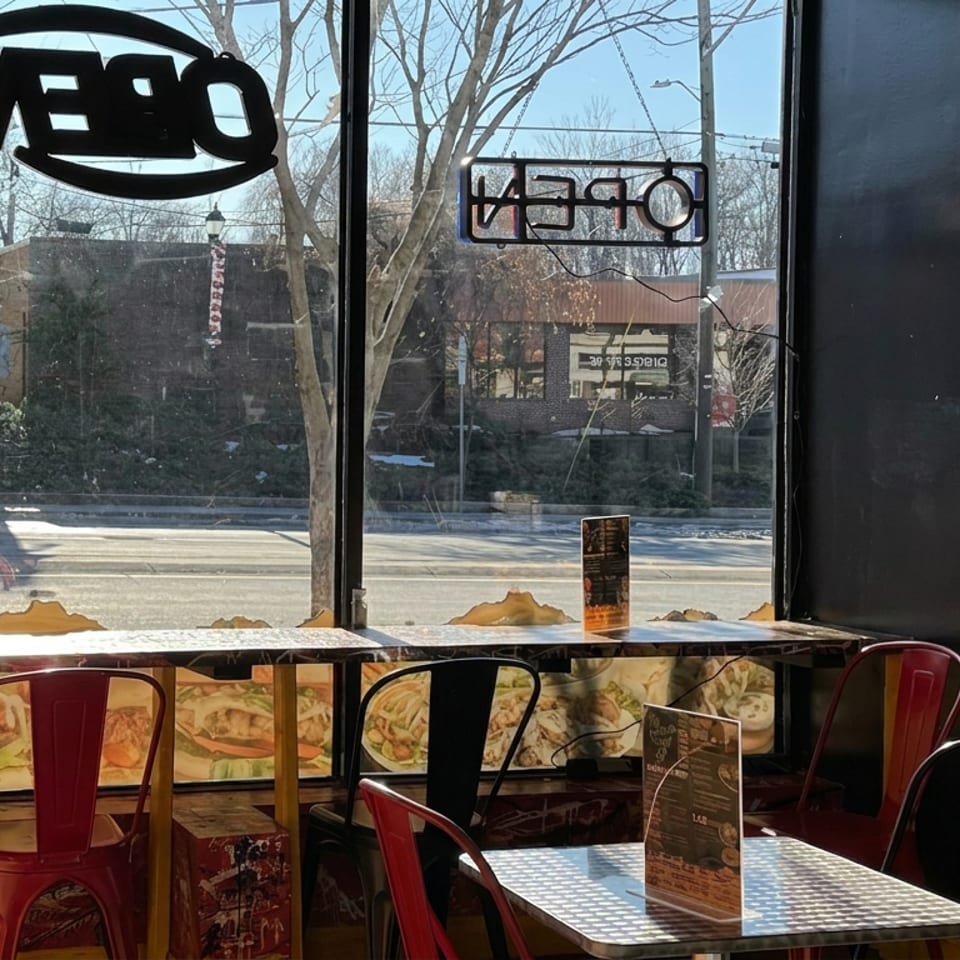 Sunlit restaurant interior with red and black metal chairs, checkered tables, window signs, menus, and a quiet street outside.