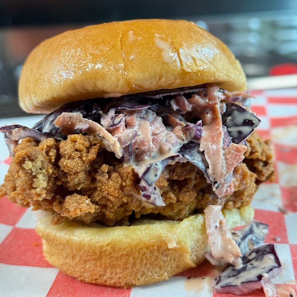 A crispy fried chicken sandwich with creamy purple coleslaw on a toasted bun, on red and white checkered paper.