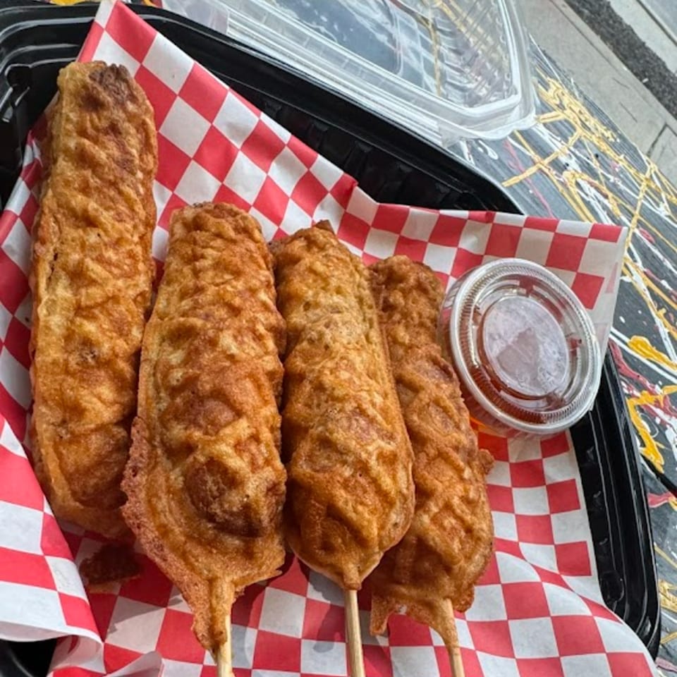 Four golden-brown Halal Hen corn dogs on a red and white checkered paper with a side of sauce.
