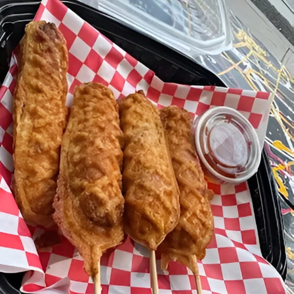 Four golden-brown Halal Hen corn dogs on sticks, served in a basket lined with red and white checkered paper.
