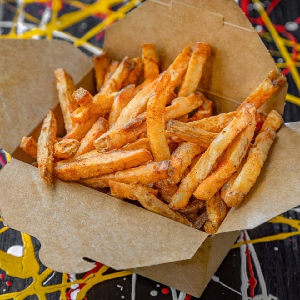 Seasoned fries in a brown paper container, on a black surface with colorful paint splatters.